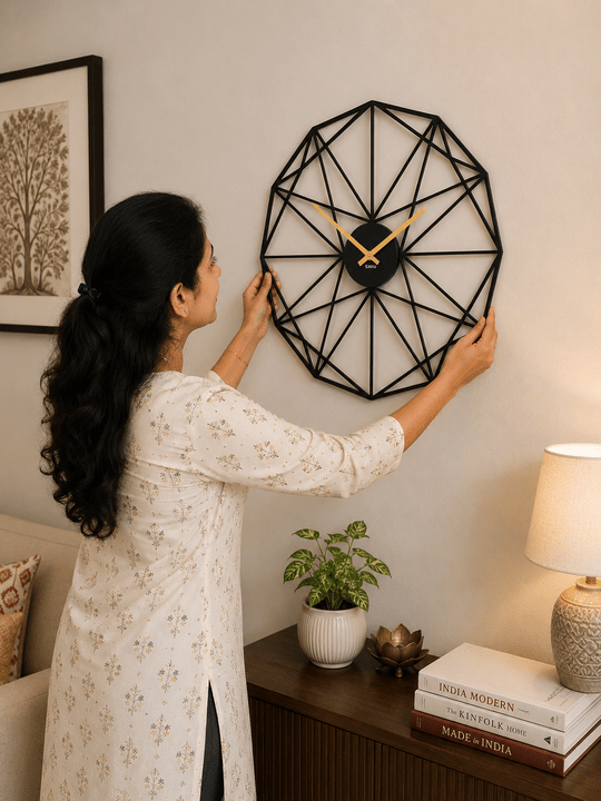 A woman mounting the large multi-faceted SHIU black polygon wall clock above a dark wood console table.