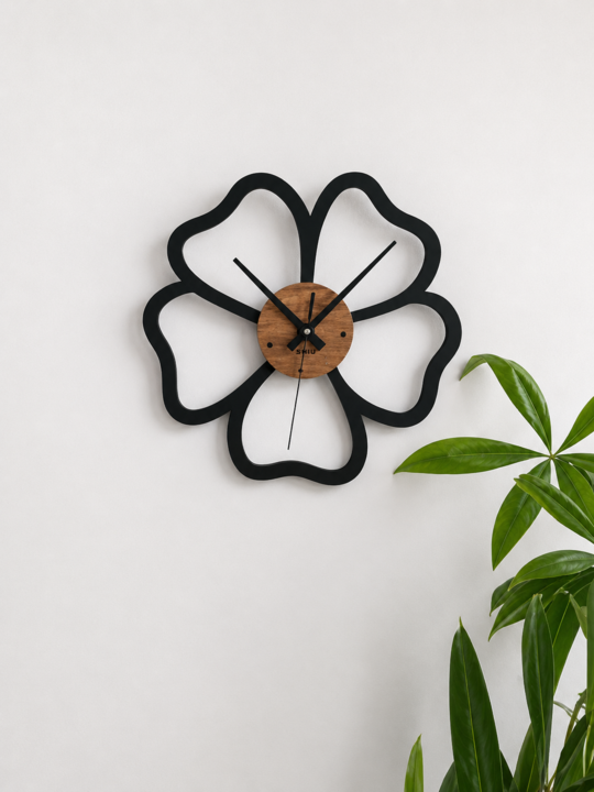 Angled view of the SHIU black open flower petal clock mounted on a white wall next to vibrant green houseplant leaves.