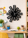 Playful SHIU abstract black word clock mounted on a white brick wall above a child's wooden study desk setup.